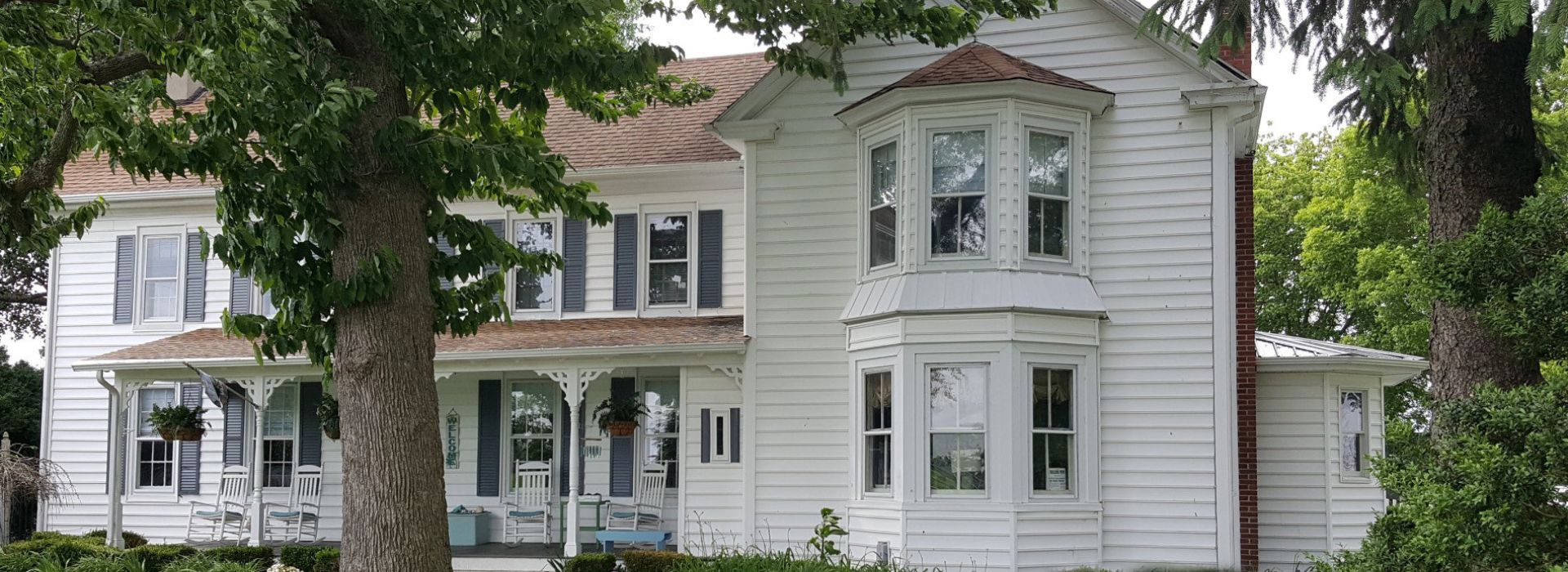 White two story historic home with rocking chairs on the front porch surrounded by lush green trees