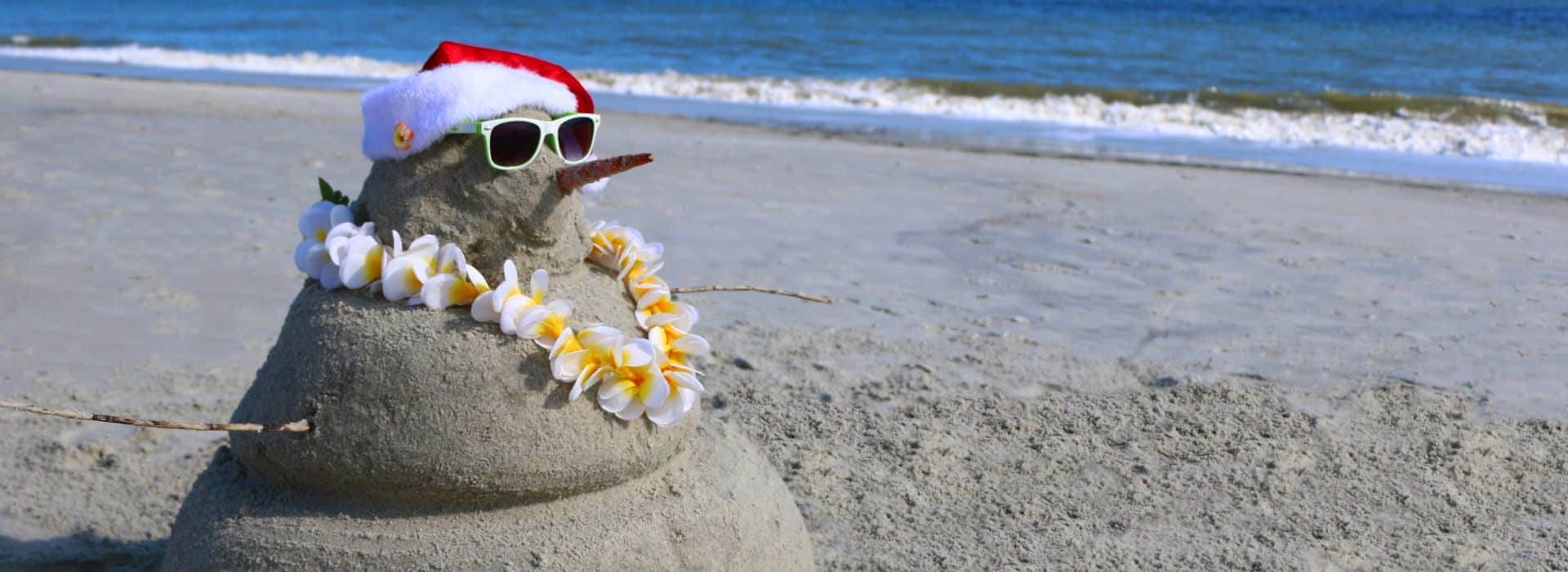 Snowman made of sand topped with a Santa hat and lei, the ocean in the background